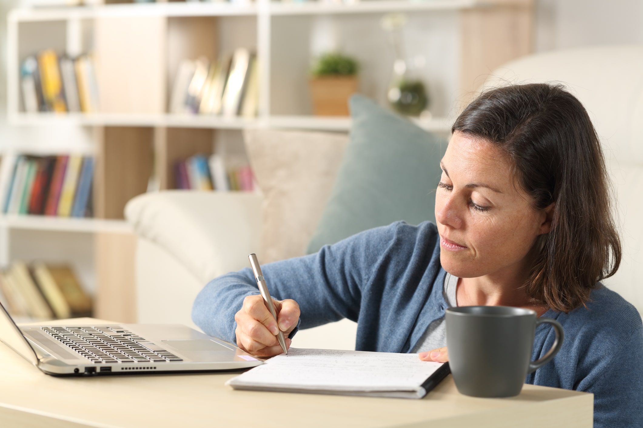 Adult woman with laptop writing on notebook at home